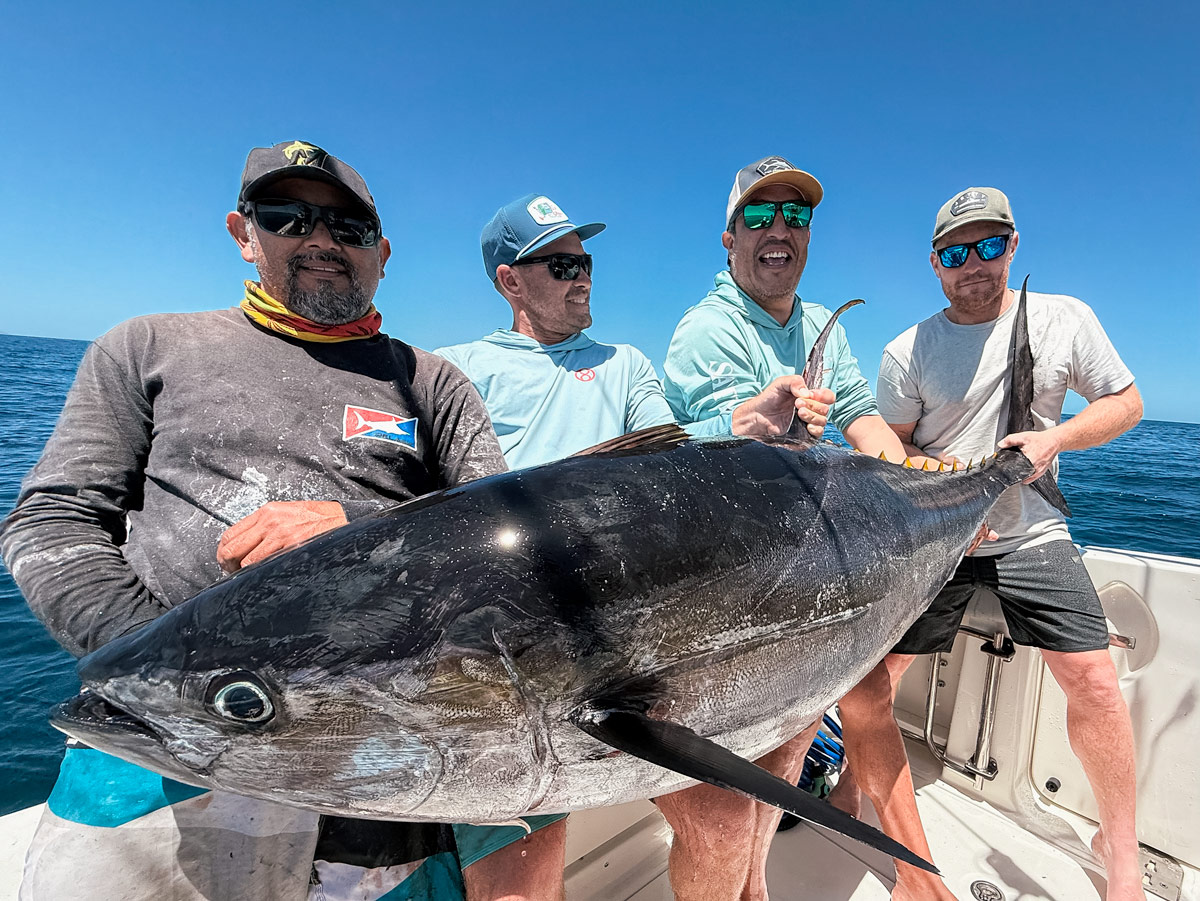 Four men holding a massive yellowfin tuna on a boat under clear blue skies.